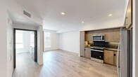 This well-designed one-bedroom, one-bathroom unit offers a thoughtful layout across a single floor, maximizing functionality within a compact footprint. The open-concept kitchen and living area serves as the heart of the space, featuring light hardwood-style flooring that flows seamlessly throughout. The kitchen is outfitted with warm wood-toned cabinetry extending to the ceiling, dark stone countertops, a dark tile backsplash, and a full suite of stainless steel appliances including a gas range, built-in microwave, dishwasher, and French door refrigerator. Recessed lighting illuminates the area efficiently.

The living area benefits from natural light provided by a glass-paneled door opening to a private outdoor balcony, accompanied by an adjacent window. The bedroom is a bright, well-proportioned room with a large window offering outdoor views and direct access to a dedicated closet equipped with built-in shelving and hanging rods. The bathroom features a wood-grain vanity with dark marble-style countertops, an undermount sink, and a backlit vanity mirror, lending a clean, contemporary feel. White walls and light flooring unify the space with a consistently modern, neutral aesthetic.