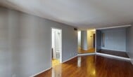 This single-floor space is designed with a straightforward, functional layout that connects several distinct areas through an open flow. The main living area is generously proportioned, featuring rich hardwood flooring that reflects light throughout the room. Gray-painted walls provide a neutral backdrop, and a textured ceiling adds subtle character overhead. A set of French doors at one end of the living area allows natural light to filter in, creating a bright and welcoming atmosphere.

The layout transitions into a secondary area with classic parquet flooring, which distinguishes it visually from the main living space. A low platform step separates these two zones, adding a modest architectural detail. The spaces are connected by a central corridor that leads to the remaining rooms.

The kitchen is compact and efficiently arranged, featuring dark brown cabinetry that extends to the ceiling for ample storage. A white coil-burner range, a full-size refrigerator, and a stainless steel sink with a tile backsplash complete the kitchen's functional setup.

A bathroom is accessible directly off the main living area, finished in light tones with basic fixtures. A walk-in closet with built-in shelving provides practical storage. HVAC vents are visible throughout, ensuring climate control across the unit.