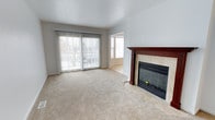 This three-floor space offers a well-organized layout designed for comfortable living. The living area, pictured here, is a generously sized room finished with neutral wall tones and plush wall-to-wall carpeting that adds warmth underfoot. A standout feature of this room is the wood-mantled fireplace, framed by tile surround and fitted with glass doors, providing both a functional heat source and a visual focal point. Natural light fills the space through a large sliding glass door fitted with vertical blinds, which also offers direct access to an outdoor area. An additional window nearby further brightens the room, giving it an open, airy feel. The transition between the carpeted living area and tiled flooring near the fireplace adds a subtle design detail that defines the space. With three floors in total, the property provides ample room for varied living, working, or storage needs. The overall aesthetic is clean and understated, with a neutral palette that serves as a versatile backdrop suitable for a range of interior styles and furnishings.