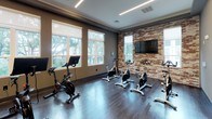 This fitness facility is thoughtfully designed to accommodate a variety of workout styles within a polished, modern environment. The space is divided into distinct zones, each serving a specific training purpose. The cycling studio is a standout feature, housing multiple stationary bikes—including Peloton-style models—arranged on rich, dark hardwood flooring. Large windows along one wall flood the room with natural light and offer views of mature trees outside, while an exposed brick accent wall adds a warm, industrial character to the space. A wall-mounted flat-screen television is positioned on the brick wall for entertainment during rides. Linear LED lighting runs across the tray ceiling, providing consistent, even illumination throughout. Adjacent to the cycling area is a functional training room equipped with stability balls, medicine balls, resistance bands, foam rollers, and a BOSU balance trainer, all neatly organized on a multi-tier storage rack. A full-length mirror spans one wall, enhancing the sense of space and allowing users to monitor their form. Sleek, glass-paneled double doors connect the studio to a broader gym area, maintaining visual openness while clearly defining each zone. The overall design balances modern aesthetics with practical functionality.