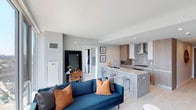 This contemporary high-rise residence is thoughtfully designed with a clean, modern aesthetic throughout. The open-concept living and kitchen area serves as the heart of the home, where light wood-toned flooring and neutral gray walls create a calm, sophisticated backdrop. The kitchen is a standout feature, boasting granite countertops with subtle veining, a large undermount sink, a gas cooktop, built-in oven, integrated microwave, and a striking stone tile backsplash. Tall cabinetry blends warm wood tones with crisp white high-gloss finishes, and a stacked in-unit washer and dryer add everyday convenience. A generously sized kitchen island with bar seating connects seamlessly to the living space. The living room is anchored by expansive floor-to-ceiling windows that frame sweeping panoramic city skyline views, flooding the space with natural light. The bedroom continues the modern design language with a bold accent wall, an open closet, and large windows offering equally impressive urban vistas. The bathroom is finished with large-format tiles, a soaking tub with an integrated showerhead, a floating wood vanity, and a wide-format undermount basin beneath a full-width mirror. Every detail reflects a refined, urban lifestyle.