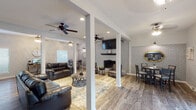 This single-floor space is designed with an open, connected layout that promotes easy flow between functional areas. The main living area is anchored by a white-painted brick accent wall housing a wood-mantled fireplace and wall-mounted television, creating a striking focal point. Multiple ceiling fans throughout ensure comfortable air circulation, while recessed lighting adds a modern, polished touch. Durable wood-look flooring runs seamlessly across the entire space.

The living area features ample seating with leather sofas and accent chairs arranged around a central coffee table, all set atop a patterned area rug. Structural columns define zones without fully enclosing them, maintaining an open and airy feel. The adjacent dining area includes a round high-top table with chairs and benefits from natural light through blinds-fitted windows, with the same white brick wall extending into this zone.

The kitchen is well-appointed with dark cabinetry, marble-look countertops, a mosaic tile backsplash, and a full suite of stainless steel appliances. A separate rear area features sliding glass doors flanked by windows, offering views of an outdoor space and allowing generous natural light to fill the room.