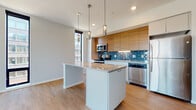 This contemporary single-floor residence showcases a thoughtfully designed open-concept layout with a clean, modern aesthetic throughout. The kitchen serves as the heart of the space, featuring warm wood cabinetry paired with crisp white lower cabinets and a central island with a light stone countertop. A striking teal subway tile backsplash adds a bold pop of color, complemented by a full suite of stainless steel appliances including a refrigerator, range, microwave, and dishwasher. Sleek pendant lighting hangs above the island, enhancing both function and style. Light-toned hardwood-style flooring flows throughout the main living and kitchen areas, contributing to the warm, cohesive feel of the space. The bedrooms are finished with plush carpet and feature expansive floor-to-ceiling windows that frame urban cityscape views, flooding the rooms with natural light. Two bathrooms provide well-appointed amenities — one includes a full soaking tub and an extended double vanity with ample cabinet storage, while the second offers a clean, efficient layout with a single vanity and mirror. Neutral wall tones and minimal detailing throughout lend the residence a refined, contemporary character well-suited to urban living.