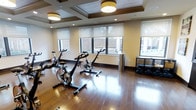 This single-floor fitness space is thoughtfully designed to support an effective workout experience. The room features rich hardwood flooring that adds warmth and durability underfoot, while a coffered ceiling with decorative molding lends an elevated architectural character uncommon in typical gym spaces. Two drum-style ceiling fixtures provide warm ambient lighting, complemented by recessed lights throughout.

Multiple stationary cycling bikes are positioned as the focal point of the room, offering a dedicated space for cardio training. Along the perimeter, a compact shelving unit houses a collection of free weights and dumbbells, providing options for strength training. A cylindrical stainless steel waste bin adds a clean, polished touch to the space.

Natural light filters in generously through several windows fitted with semi-sheer roller shades, offering views of the surrounding brick exterior and greenery outside. The shades allow residents to control light levels while maintaining a bright, energizing atmosphere. Overall, the space balances functional fitness amenities with refined design details, creating an inviting and well-lit environment for daily exercise.