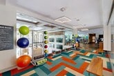 Located at 3400 Davie Rd in Davie, Florida, this single-floor fitness facility offers a generously sized, thoughtfully designed space dedicated to health and wellness. The layout encompasses two distinct exercise rooms, connecting hallways, and a dedicated office, all flowing seamlessly together to create a comprehensive fitness environment.

The cardio and functional training room is immediately eye-catching, featuring a bold, geometric patterned carpet in vibrant teal, orange, and cream tones. This room is equipped with treadmills, ellipticals, stationary bikes, and water rowers, alongside a dedicated functional training area stocked with stability balls, medicine balls, TRX suspension systems, and balance trainers. Floor-to-ceiling glass partitions allow natural light to pour through while maintaining an open, airy feel. Square pendant light fixtures add a modern architectural touch to the bright white ceilings.

The strength training room transitions to durable rubberized flooring and is outfitted with an extensive array of free weights, dumbbells, cable machines, Smith machines, and selectorized weight equipment. Full-length mirrors line the walls, amplifying the sense of space. Large windows offer views of the surrounding greenery, and teal accent walls provide a consistent, energizing design theme throughout.

A charming pet grooming spa rounds out the facility, featuring tiled walls, wood cabinetry, and a self-service dog wash station — a unique and practical amenity.