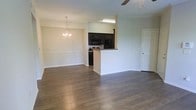 This single-floor space features an open-concept layout that seamlessly connects the living, dining, and kitchen areas. The rich, dark hardwood flooring extends throughout, providing a warm and cohesive foundation. The dining area is accentuated by an elegant chandelier, adding a refined touch, while a ceiling fan serves the adjoining living space, offering both style and functionality. The kitchen is defined by a peninsula with granite countertops, creating a natural divide without sacrificing the open feel. Dark cabinetry contrasts sharply against the light walls, and black appliances — including a built-in microwave, refrigerator, and range — complete the sleek, modern aesthetic. Neutral, light-toned walls throughout keep the space feeling bright and airy. A white paneled door provides access to an additional room or storage area. The overall ambiance is clean and contemporary, balancing practical design with inviting details that make the space feel both functional and welcoming.