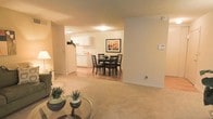 This single-floor residence offers an open-concept layout that seamlessly connects the main living areas, creating a cohesive and functional flow throughout the home. The living area is finished with soft, neutral-toned carpeting, providing a comfortable and inviting atmosphere. A glass-topped coffee table and tasteful greenery accent the space, adding subtle decorative touches.

The living area opens directly into a dedicated dining space, which features hardwood-style flooring and accommodates a dark-finished dining table and chair set, complemented by framed wall art that adds a warm focal point to the room. This transition from carpet to hard flooring clearly delineates the living and dining zones while maintaining an airy, connected feel.

Adjacent to the dining area, the kitchen is outfitted with crisp white cabinetry, light countertops, and full-sized appliances, including a refrigerator. The cabinetry extends to the upper walls, offering ample storage. Overhead lighting in both the kitchen and dining areas ensures the space remains bright and well-lit. The overall color palette throughout is warm and neutral, with beige and white tones contributing to a clean, welcoming ambiance suited to everyday living.