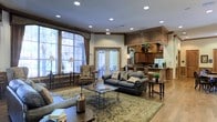 This single-floor space presents a thoughtfully designed interior that blends classic elegance with comfortable functionality. The main lounge area serves as the centerpiece, anchored by a pair of dark leather sofas arranged around a glass-top coffee table atop a large ornate area rug. Complementary seating, including a patterned upholstered armchair, adds to the welcoming arrangement.

Natural light pours in generously through oversized windows dressed with decorative drapes, as well as a set of French doors that open to an outdoor seating area, providing a strong visual connection to the exterior. Recessed lighting in the ceiling supplements the ambient glow from traditional floor lamps, ensuring the space remains well-lit throughout.

Rich wood tones are a defining characteristic, featured prominently in the cabinetry of the adjacent kitchen or service area, which includes dark wood upper cabinets, a raised bar counter, and appliances. A bold wood architectural element frames the kitchen area, adding a sense of grandeur. The overall ambiance is warm, inviting, and traditionally styled, suitable for both socializing and relaxed gatherings.