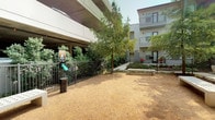 This outdoor dog park is a fully enclosed, ground-level space designed to provide a safe and functional environment for pets and their owners. Access is controlled through a sturdy metal gate equipped with a keypad lock, ensuring the area remains secure for authorized users only.

The park's ground cover consists of packed gravel and decomposed granite, providing a durable, low-maintenance surface well-suited for active use. Several mature trees are distributed throughout the space, offering natural shade that helps moderate temperatures during warmer conditions. Concrete benches are positioned throughout the area, giving owners comfortable places to sit and supervise their pets.

The perimeter is defined by a tall metal fence that maintains visibility while containing pets safely within the space. Waste disposal stations are conveniently located near the entrance, promoting cleanliness and responsible pet ownership. The park is bordered by a multi-story parking structure on one side and residential buildings on others, situating it within a larger urban community setting. Landscaped shrub beds along the fencing add greenery and visual interest to the overall environment.
