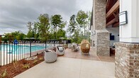 Located at 1821 Chatham Flats Dr in Apex, North Carolina, this outdoor amenity space is a thoughtfully designed community gathering area that combines resort-style living with functional outdoor entertaining. At its centerpiece is a spacious, crystal-clear swimming pool enclosed by sleek black iron fencing, surrounded by concrete pool decking outfitted with lounge chairs and patio umbrellas for relaxation.

Adjacent to the pool is a covered pavilion that serves as the social hub of the space. Soaring exposed wood beam ceilings and a vaulted roofline lend warmth and architectural character to the structure. Inside the pavilion, shiplap-style white walls contrast beautifully with the rich wood tones overhead. The space is equipped with mounted flat-screen televisions, a built-in outdoor kitchen counter with sink, and a large dining table with rope-woven chairs, making it ideal for gatherings. A prominent brick fireplace anchors one side of the pavilion, adding ambiance and year-round usability.

The surrounding patio areas feature a mix of stamped concrete and wood-plank decking, furnished with woven lounge chairs and rattan accent seating. Mature trees and professionally landscaped garden beds frame the perimeter, providing natural screening and a serene backdrop. The overall atmosphere is polished, inviting, and well-suited for both casual relaxation and social events.