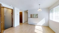 This three-floor unit at 174 A offers a clean, functional interior with a straightforward layout. The main living area is finished with light-toned hardwood-style flooring that reflects natural sunlight, creating a bright and airy atmosphere throughout the space. White walls provide a neutral backdrop, while a textured ceiling adds subtle visual character overhead.

A pendant light fixture hangs above the dining area, which connects to the kitchen through a pass-through window — a practical architectural detail that maintains an open feel between the two spaces. Baseboard heating units are positioned along the walls, ensuring efficient warmth throughout the colder months. The hallway leads further into the unit, where a warm-toned wood door anchors the corridor and hints at the additional rooms beyond.

Natural light filters in through the pass-through window, offering a glimpse of greenery outside and enhancing the overall sense of openness. The multi-floor configuration provides distinct separation between living, sleeping, and utility areas, making for a well-organized and versatile space suited to a variety of lifestyles.