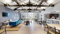 Located at 7820 Lexington Ave in Cleveland, Ohio, this single-floor community space offers a generous, open layout designed for both functionality and comfort. The space is defined by soaring ceilings with exposed industrial ductwork, lending it a loft-like character that feels both modern and inviting.

The well-appointed kitchen is a standout feature, showcasing sleek wood-grain cabinetry, open shelving stocked with décor and essentials, a marble-look quartz island with bar seating, stainless steel appliances including a full-size range and refrigerator, and a subway tile backsplash. A row of pendant lights illuminates the island, enhancing the kitchen's contemporary aesthetic against dark hardwood-style flooring.

The dining area offers multiple seating configurations, including a round wooden table with cushioned chairs and a rectangular table with vibrant green accent chairs. Large floor-to-ceiling windows flood the dining space with natural light and provide views of the surrounding greenery outside. Framed artwork adorns the neutral-toned walls, adding a polished, curated feel throughout.

The overall ambiance is clean, modern, and welcoming — a well-designed communal space suited for gathering, dining, and everyday use.
