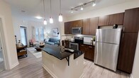 This single-floor unit offers an efficient and thoughtfully designed open-concept layout that connects the kitchen and living area seamlessly. The kitchen serves as a focal point of the space, featuring rich dark wood cabinetry paired with sleek black granite countertops that extend onto a central island with an integrated sink and seating overhang — ideal for casual dining or entertaining. A full suite of stainless steel appliances, including a refrigerator, range, and built-in microwave, adds a polished, contemporary touch. Pendant lighting above the island, complemented by track lighting overhead, ensures the kitchen is well-lit and visually striking. Light wood-style flooring runs throughout, contributing to a bright and cohesive aesthetic. The open living area is visible from the kitchen and benefits from natural light filtering through a window dressed with classic shutters. White walls keep the space feeling airy and open. The overall ambiance is modern and clean, combining functional design with stylish finishes suited for comfortable everyday living.