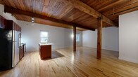 Located at 219 E Bank St in Petersburg, VA, this one-bedroom, one-bathroom unit blends historic character with modern updates across a well-proportioned single floor. The open-concept kitchen and living area is the centerpiece of the space, featuring soaring wood-plank ceilings supported by exposed original timber beams and posts that speak to the building's storied past. Light hardwood flooring runs throughout, complementing the warm tones of the ceiling and structural elements. The kitchen is well-appointed with rich mahogany-toned cabinetry, a white quartz island with an undermount sink, and a full suite of stainless steel appliances, including a refrigerator, range, and built-in microwave. Track lighting illuminates the space effectively. The bedroom offers a comfortable footprint with a continuation of the hardwood floors, a wood-plank ceiling, a window for natural light, and a striking exposed brick accent wall that adds texture and historic charm. The bathroom features classic penny-tile flooring, a single-vanity with matching cabinetry, and a tub-shower combination. Additional spaces include a dedicated laundry area, a closet, and a utility room, providing practical storage and functionality throughout.