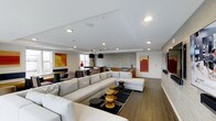 This single-floor space showcases a thoughtfully designed open-plan layout that seamlessly integrates a spacious living area, a dining zone, and a kitchenette into one cohesive environment. The living area is anchored by a generous white sectional sofa and chaise, paired with a bold black-and-white area rug and a collection of sculptural wooden accent tables that add warmth and visual interest. Recessed ceiling lighting throughout ensures the space is well-illuminated, complemented by natural light filtering in through multiple windows. The floors are finished in rich hardwood-tone planks that run consistently across the entire space, lending a sense of warmth and continuity. Vibrant abstract artwork in warm amber and red tones punctuates the neutral white walls, adding personality and color. A large flat-screen television is mounted on a paneled feature wall, creating a dedicated entertainment focal point. Toward the rear of the space, a dining area with dark furnishings and pendant lighting transitions naturally into a compact kitchenette finished with a textured tile backsplash. The overall ambiance is modern, sophisticated, and inviting, with a clean aesthetic that balances comfort and contemporary style throughout.