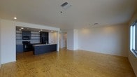 This single-floor space showcases a thoughtfully designed open-concept layout that seamlessly connects the kitchen and living areas. The kitchen is a standout feature, defined by sleek dark cabinetry that extends to the ceiling, open shelving along one wall for functional storage display, and a navy-toned island with an integrated sink. The island serves as a natural dividing element between the kitchen and the expansive living area, making it ideal for both meal preparation and casual gathering.

The flooring throughout is a distinctive natural plywood, offering warm, pronounced wood grain patterns that add an industrial-yet-organic character to the space. The smooth white walls and flat white ceilings create a clean, neutral backdrop that complements the contrast of the dark kitchen elements. Recessed lighting in the kitchen area provides focused task illumination, while ceiling-mounted HVAC vents ensure climate control throughout.

The living area is generously proportioned and open, providing flexibility for various furniture arrangements. A hallway visible in the background suggests access to additional rooms or an entry, contributing to the overall functional flow of this modern, minimalist space.