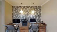 This single-floor office space is thoughtfully designed to balance functionality with refined aesthetics. The most striking architectural feature is the floor-to-ceiling mosaic tile accent wall, composed of earthy brown, beige, and gray tones arranged in a brick-style pattern, creating a rich and textured focal point that anchors the room.

Two wooden desks are arranged side by side against this feature wall, each equipped with an Apple iMac workstation, providing a dual-workstation setup ideal for collaborative or parallel workflows. The desks offer generous surface area and built-in storage drawers, while ergonomic mesh chairs ensure comfort during extended work sessions. A shared multifunction printer is centrally positioned between the two workstations for convenient access.

Overhead, two modern pendant lights with frosted globe-shaped shades hang from the ceiling, casting a warm, ambient glow that complements the mosaic wall and the natural wood tones of the furniture. Crown molding along the ceiling adds a polished, finished look to the overall design.

The space exudes a professional yet inviting atmosphere, combining practical office infrastructure with deliberate design choices that elevate it beyond a standard workspace. The cohesive color palette of warm neutrals and wood tones throughout creates a focused and comfortable environment.