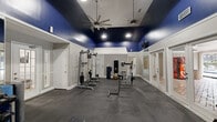 This fitness center is a thoughtfully designed workout facility that offers a full range of exercise equipment within a compact yet efficient single-room layout. The space is immediately distinguished by its bold navy-painted upper walls and ceiling, which contrast sharply with the light gray lower walls accented by white chair rail molding, creating a dynamic and energizing atmosphere. The elevated ceiling height adds a sense of openness, and a large ceiling fan keeps the space comfortable during workouts.

The rubber flooring provides a durable, impact-absorbing surface suited for both strength training and cardio activities. A large wall of floor-to-ceiling mirrors along one side visually expands the room while allowing users to monitor their form. The cardio section features treadmills, an elliptical, and a stationary bike, while the strength training area includes a multi-station cable machine, a weight bench, dumbbells, and additional resistance equipment.

Recessed lighting and a wall-mounted television enhance functionality. A standout feature is the set of French doors that open directly to an outdoor pool and patio area, flooding the space with natural light and providing convenient access to outdoor amenities. The overall ambiance is polished and motivating, making it a practical and well-appointed fitness destination.