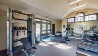 This fitness center offers a comprehensive workout environment housed within an architecturally appealing single-floor facility. The space features soaring vaulted ceilings with a prominent arched window and multiple transom-style windows that flood the interior with natural light, creating an open and energizing atmosphere. Durable rubber flooring spans the entire space, providing both safety and comfort underfoot.

The cardio zone is well-appointed with multiple Matrix treadmills and elliptical machines, all positioned to take advantage of the natural light streaming in from large glass doors and windows that offer views of the surrounding wooded landscape. A dedicated spinning area features stationary cycling bikes, complemented by wall-to-wall mirrors that visually expand the space and allow users to monitor their form during workouts.

The strength training area is equipped with a multi-functional cable machine for a variety of resistance exercises, along with a full rack of dumbbells in graduated weights, stability balls, and a folding exercise mat. An adjustable weight bench rounds out the free weight section. A water station is conveniently accessible near the entrance, and large glass doors provide direct access to an outdoor pool area, seamlessly connecting indoor and outdoor amenities. A wall-mounted television adds entertainment options for cardio sessions.