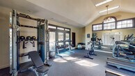 This fitness center offers a comprehensive workout environment housed within an architecturally appealing single-floor facility. The space features soaring vaulted ceilings with a prominent arched window and multiple transom-style windows that flood the interior with natural light, creating an open and energizing atmosphere. Durable rubber flooring spans the entire space, providing both safety and comfort underfoot.

The cardio zone is well-appointed with multiple Matrix treadmills and elliptical machines, all positioned to take advantage of the natural light streaming in from large glass doors and windows that offer views of the surrounding wooded landscape. A dedicated spinning area features stationary cycling bikes, complemented by wall-to-wall mirrors that visually expand the space and allow users to monitor their form during workouts.

The strength training area is equipped with a multi-functional cable machine for a variety of resistance exercises, along with a full rack of dumbbells in graduated weights, stability balls, and a folding exercise mat. An adjustable weight bench rounds out the free weight section. A water station is conveniently accessible near the entrance, and large glass doors provide direct access to an outdoor pool area, seamlessly connecting indoor and outdoor amenities. A wall-mounted television adds entertainment options for cardio sessions.