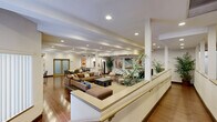 Located at 125 W Olive Ave in Monrovia, California, this single-floor space offers a generously sized, open-plan interior designed for comfort and functionality. The main lounge area is a standout feature, showcasing rich hardwood-style flooring, a distinctive coffered ceiling with extensive recessed lighting, and structural columns that lend an architectural character to the room. Large vertical-blind windows along the perimeter allow natural light to filter throughout, creating a bright and welcoming ambiance. The lounge is thoughtfully laid out with multiple seating arrangements, including large sectional sofas, accent chairs, and coffee tables, all anchored by a neutral area rug. A large flat-screen television serves as a focal point in one seating zone, while vibrant wall art adds color and personality to the space. Lush indoor plants are strategically placed throughout, enhancing the fresh, inviting atmosphere. The kitchen is sleek and modern, featuring white cabinetry, a dark subway tile backsplash, stainless steel appliances including a range with a wall-mounted hood and a full-size refrigerator, and a granite-topped breakfast bar with bar seating. A dedicated dining area with table and chairs is visible adjacent to the kitchen, completing this well-appointed communal space.