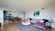 This single-floor residence offers a thoughtfully designed open-plan layout that integrates the living, dining, and kitchen areas into one cohesive, light-filled space. The interior is finished with clean white walls and smooth ceilings that amplify the sense of openness, while warm light wood flooring runs throughout, adding a natural and inviting contrast.

The living area is anchored by a comfortable grey sofa accented with colorful throw pillows and a bold geometric black-and-white area rug, lending the space a contemporary edge. A striking large-scale abstract artwork in teal and green tones serves as a focal point on the main wall, reflecting an artistic sensibility. A sleek circular white coffee table completes the seating arrangement.

The dining area transitions naturally from the living space, featuring a round dining table paired with warm wood-toned chairs — a practical yet stylish setup. Adjacent to the dining area, the kitchen is finished in crisp white cabinetry with pendant lighting overhead, offering a clean and functional cooking environment with open visibility to the rest of the space.

Natural light fills the interior through well-positioned windows, enhancing the airy atmosphere. Ceiling-mounted light fixtures and visible ventilation elements point to a well-equipped, move-in ready home. Overall, the space strikes a balance between modern minimalism and expressive, curated design.