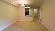 This single-floor residential unit offers a well-organized and functional layout designed for comfortable everyday living. The main living area is generously proportioned, featuring soft wall-to-wall carpeting in a warm beige tone that creates an inviting atmosphere. Crown molding lines the ceiling, adding a refined architectural detail, while a pendant light fixture serves as a central focal point overhead.

The kitchen is positioned adjacent to the living space and features a pass-through counter with a built-in sink, providing both a visual connection and practical convenience between the two areas. Cabinetry in a light wood finish lines the upper kitchen walls, and a black refrigerator anchors one end of the kitchen. Recessed under-cabinet lighting adds functional illumination to the workspace.

A hallway extending from the main living area leads to additional rooms, flanked by multiple doors — suggesting several private spaces such as bedrooms and a bathroom. The hallway is finished with consistent neutral-toned walls and benefits from recessed overhead lighting, maintaining the cohesive, clean aesthetic found throughout the unit.

Overall, the space presents a neutral, move-in-ready palette with consistent finishes, making it highly adaptable for a variety of furnishing styles and personal touches.