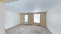 This single-story space presents a clean, open layout designed for versatile use. The main room is finished with warm beige walls and white trim that create a neutral, welcoming atmosphere throughout. Soft, wall-to-wall carpeting in a light tan tone covers the floor, adding comfort underfoot and contributing to the room's understated aesthetic. The smooth white ceiling complements the light color palette, enhancing the sense of openness within the space. Natural light enters generously through two windows fitted with white blinds, as well as a glass-paneled door that provides both illumination and outdoor access. The windows offer views of surrounding greenery, further brightening the interior. Electrical outlets are conveniently positioned along the walls, and baseboard heating runs along the perimeter, ensuring practical functionality. The overall ambiance is clean, airy, and move-in ready, offering a blank canvas suitable for a variety of living or working arrangements. The straightforward single-floor layout adds to the space's accessibility and ease of use.