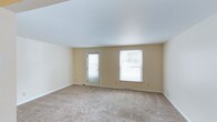 This single-story space presents a clean, open layout designed for versatile use. The main room is finished with warm beige walls and white trim that create a neutral, welcoming atmosphere throughout. Soft, wall-to-wall carpeting in a light tan tone covers the floor, adding comfort underfoot and contributing to the room's understated aesthetic. The smooth white ceiling complements the light color palette, enhancing the sense of openness within the space. Natural light enters generously through two windows fitted with white blinds, as well as a glass-paneled door that provides both illumination and outdoor access. The windows offer views of surrounding greenery, further brightening the interior. Electrical outlets are conveniently positioned along the walls, and baseboard heating runs along the perimeter, ensuring practical functionality. The overall ambiance is clean, airy, and move-in ready, offering a blank canvas suitable for a variety of living or working arrangements. The straightforward single-floor layout adds to the space's accessibility and ease of use.