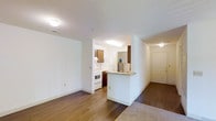 This single-floor unit offers a practical and well-organized layout designed for comfortable everyday living. The open-concept living and dining area is finished with warm hardwood-style flooring that transitions smoothly into plush carpeting, creating distinct zones within the shared space. Neutral white walls and flush-mount ceiling lighting keep the interior feeling bright and clean throughout.

The kitchen is positioned adjacent to the living area and is accessible via a half-wall peninsula featuring a laminate countertop, which serves as both a breakfast bar and a natural divider between the kitchen and main living space. The kitchen is equipped with wood-toned cabinetry, a white electric range, a refrigerator, and additional appliances, offering a fully functional cooking environment. A secondary appliance, visible near the back wall, adds further utility.

A hallway leads from the living area toward additional rooms, with a doorway visible that connects to what appears to be a bedroom or secondary space, also carpeted. A closet with standard white doors is conveniently located near the entryway, providing ample storage. The front entry door is solid and well-positioned to create a clear transition from the exterior into the living space. The overall ambiance is clean, neutral, and move-in ready.