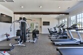 Located in Bloomingdale, Georgia, this single-story fitness center offers a thoughtfully designed workout space that balances functionality with an inviting atmosphere. The interior is finished with neutral white walls, crown molding, and distinctive dark wood exposed beams running across the ceiling, adding a warm architectural contrast to the clean, modern aesthetic. Recessed lighting throughout ensures bright, even illumination across the entire space.

The fitness area is well-appointed with multiple treadmills and an elliptical machine lined along a wall of large, multi-pane windows that flood the room with natural light and provide views of the surrounding outdoor environment. A multi-station cable weight machine and a flat workout bench occupy a dedicated strength training zone, complemented by medicine balls for functional training exercises. A wall-mounted flat-screen television is positioned for easy viewing during workouts.

The flooring consists of durable, speckled rubber gym flooring suitable for high-traffic fitness use. A glass double-door entry with an exit sign provides convenient access to the exterior. The overall ambiance is clean, modern, and practical, creating a welcoming environment well-suited for a comprehensive fitness routine.