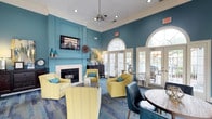 This single-floor clubhouse is thoughtfully designed to serve as a welcoming social hub, combining comfort and style in a cohesive interior. The space is defined by its bold teal accent walls, crisp white crown molding, and soaring ceilings that lend an open, airy feel throughout. Warm hardwood flooring transitions into vibrant multi-toned carpeting, delineating the seating and gathering areas with visual clarity.

The main lounge area is anchored by a classic fireplace topped with a wall-mounted flat-screen television, creating an inviting focal point. Plush yellow armchairs and a tufted white sofa are arranged to encourage relaxed conversation, accented with teal and chartreuse throw pillows that complement the room's color story. A round wooden dining table flanked by navy blue leather chairs provides a more structured setting for gatherings.

Abundant natural light pours in through a series of arched French doors fitted with transom windows, offering views of and access to an outdoor patio area. Recessed lighting and a ceiling fan ensure year-round comfort. Decorative touches, including wall art, table lamps atop a dark wood credenza, and coastal-inspired accents, complete the polished yet relaxed atmosphere. A hallway with a decorative round mirror connects to additional service areas, enhancing the overall functionality of the space.