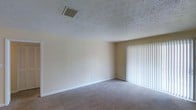 This single-floor space presents a straightforward and functional layout characterized by clean lines and neutral design choices. The main living area is finished with wall-to-wall carpet in a warm beige tone, providing a comfortable underfoot feel throughout. The walls are painted in a soft, neutral cream hue that keeps the space feeling bright and open, while the textured popcorn ceiling adds a classic residential touch overhead. A central HVAC vent is integrated into the ceiling, ensuring efficient climate control throughout the interior. Natural light enters the room through a sliding glass door or large window fitted with white vertical blinds, which can be adjusted to control light and privacy as needed. White baseboards run along the perimeter of the room, offering a clean contrast against the carpeted floor. The overall ambiance of the space is calm and understated, making it a versatile canvas for a variety of furnishing styles and décor preferences. The single-floor layout ensures easy accessibility and straightforward navigation throughout, making it a practical and welcoming environment.