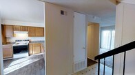 This two-story space presents a practical and functional layout across its two levels. The main level features a kitchen area equipped with warm oak cabinetry and laminate countertops, paired with durable wood-look flooring that adds a cohesive feel. A textured ceiling runs throughout, contributing to the overall character of the interior.

A central hallway connects the kitchen to the adjacent living area, where a vertical blind-covered window allows natural light to filter in. The flooring transitions from tile with a subtle diamond-pattern detail near the entryway to wood-look planks in the living space, defining each zone clearly. A white interior door with a brass-toned handle provides access to an additional room or closet, while HVAC vents positioned both above and below indicate a well-integrated climate control system throughout.

The staircase, framed with a dark metal railing, leads to the upper level, suggesting additional living or sleeping quarters above. The overall aesthetic is clean and neutral, with beige and white tones dominating the walls, making the space adaptable for a variety of furnishing styles and uses.