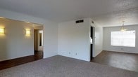 This single-story space presents a straightforward and functional layout designed for comfortable everyday living. The main living area is finished with neutral-toned carpet underfoot, providing a soft and practical surface throughout the central space. Crisp white walls create a clean, bright backdrop, lending the interior a simple and versatile aesthetic that can accommodate a variety of furnishings and décor styles.

Overhead, a standard flat ceiling houses centrally placed HVAC vents, ensuring climate control is distributed throughout the space. An open doorway on the left side of the main room leads into a connecting hallway, where wood-look flooring transitions the space into what appears to be a corridor accessing additional rooms. This hallway glimpse hints at further rooms beyond the main living area, suggesting a reasonably connected and navigable floor plan.

To the right, a doorway opens into what appears to be a secondary room or utility space, partially visible and featuring its own lighting and access point. The overall ambiance of the space is neutral and unadorned, making it a blank canvas well-suited for personalization. The layout emphasizes practicality, with a clean flow between areas and consistent finishes throughout.