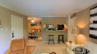 This single-floor residence offers a thoughtfully designed open-concept layout that connects the main living areas with ease and functionality. The living room is warm and inviting, featuring plush carpeting, crown molding, and a textured ceiling that adds subtle character throughout the space. A pass-through kitchen bar with seating for two serves as the central hub of the home, bridging the living area with the kitchen. The kitchen is equipped with dark cabinetry and a full suite of appliances, including a black refrigerator, and benefits from a window that draws in natural light. The dining area flows naturally from the living room, complete with a ceiling fan and ample space for a full dining set — ideal for both casual meals and entertaining. The warm, neutral color palette used throughout creates a cohesive and comfortable ambiance, while the consistent carpeting underfoot adds to the sense of continuity across the open floor plan. Crown molding details along the ceilings lend a refined touch to the overall aesthetic, making this a practical yet polished living space.