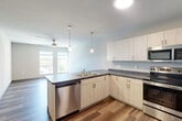 This contemporary 2-bedroom, 1-bathroom residence is designed with a clean, modern aesthetic and functional layout across a single floor. The open-concept kitchen and living area serves as the heart of the home, featuring light wood-tone cabinetry, dark countertops, and a full suite of stainless steel appliances including a refrigerator, electric range, built-in microwave, and dishwasher. Pendant lighting hangs above the kitchen peninsula, which includes a double sink and provides additional counter space. Wood-look plank flooring runs throughout the main living areas, adding warmth and visual continuity. A ceiling fan in the living area enhances comfort, while large windows invite natural light and offer outdoor views. The primary bedroom is generously sized, finished with plush carpet, a ceiling fan, and windows that frame exterior views. The second bedroom features wood-look flooring and neutral walls, offering versatile use. The shared bathroom is well-appointed with a floating-style vanity, white countertop, large mirror with contemporary lighting, and matching wood-tone cabinetry. A dedicated closet provides added storage. The unit's neutral color palette and consistent modern finishes create a cohesive, move-in ready living environment.