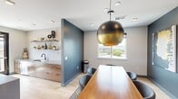 Located at 281 Willow St in Fort Collins, Colorado, this thoughtfully designed single-floor space blends modern aesthetics with functional versatility. Light hardwood flooring runs throughout, complementing a neutral gray and slate-blue color palette that lends the room a cohesive, contemporary feel.

The lounge area is anchored by a striking vertical-slat accent wall in deep charcoal, against which a large flat-screen TV and a modern media console are centered. A cognac leather sectional sofa faces the display, positioned to take full advantage of the expansive windows that frame elevated outdoor views of the Fort Collins skyline and surrounding greenery.

Transitioning into the dining zone, a substantial wood-topped conference-style table is paired with upholstered modern chairs and presided over by a bold globe pendant light finished in brushed gold and black. An eye-catching triptych of large-scale abstract artwork adorns the slate-blue accent wall, adding a gallery-quality artistic statement to the space.

The kitchenette area features warm wood cabinetry, a white countertop with an undermount sink, open floating shelves styled with decorative objects, and a concrete-topped kitchen island with bar seating. A gold and black pendant cluster light adds an elegant focal point above. Floor-to-ceiling windows and a glass door open to an outdoor balcony, flooding the space with natural light.