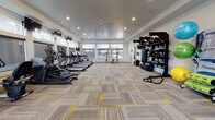 This modern fitness center offers a spacious, single-floor layout designed to accommodate a comprehensive range of workout routines. The room is finished with a stylish patterned carpet featuring gray and yellow accents, complementing the clean white walls and recessed lighting overhead. Ceiling fans are positioned throughout the space to ensure comfortable airflow during workouts.

Along one side of the facility, a row of treadmills and elliptical machines is neatly arranged, providing ample cardio options. Large windows line the perimeter, flooding the interior with natural light and offering views of the surrounding outdoor environment. A multi-station cable and pulley machine is centrally positioned near the windows, supporting a variety of strength training exercises.

The strength and functional training area is well-stocked with a full rack of dumbbells, a multi-purpose power rack, TRX suspension trainers, battle ropes, and a dedicated storage unit housing medicine balls, kettlebells, and resistance bands. An adjustable weight bench adds further versatility. A colorful ball rack displays stability balls and foam rollers, supporting flexibility and core training.

Wall-mounted flat-screen televisions are positioned throughout the space for entertainment during workouts. A glass entry door provides direct outdoor access, contributing to the open, airy ambiance of this comprehensively outfitted fitness facility.