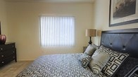 The bedroom is a well-appointed space designed with comfort and simplicity in mind. The room is finished with smooth, neutral-toned walls that create a calm and inviting atmosphere. A large window dressed with vertical blinds serves as the focal point of the main wall, allowing natural light to filter softly into the room while offering privacy control. The window's generous size contributes to a bright and airy feel throughout the space. The room comfortably accommodates a full-sized bed positioned against one wall, complete with a upholstered headboard in a sleek charcoal tone. Coordinating patterned bedding in grey and blue geometric tones adds a modern, stylish touch. A bedside table with a warm-toned table lamp sits adjacent to the bed, providing functional ambient lighting. The neutral carpeted flooring ties the space together, adding warmth underfoot. Overall, the bedroom offers a clean, understated aesthetic with thoughtful details that balance practicality and style, making it a restful retreat within the home.