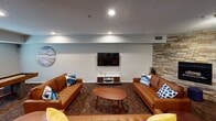 This single-floor communal space is thoughtfully designed to accommodate a variety of social and recreational activities. The centerpiece of the lounge area is a dramatic floor-to-ceiling stacked stone accent wall housing a fireplace with a marble-style hearth, creating a warm and inviting focal point. Two cognac leather sofas, accented with blue throw pillows, flank an oval wooden coffee table, while a wall-mounted flat-screen TV and a sleek media console complete the entertainment setup.

Adjacent to the lounge, a dedicated casual dining area features round white tables paired with modern teal chairs, set beneath hexagonal wall art that adds a playful, contemporary touch. Natural light filters in through nearby windows, brightening this corner of the space.

A full kitchen with white shaker cabinetry, stainless steel appliances—including a refrigerator, gas range, microwave, and dishwasher—and dark granite countertops provides ample functionality for hosting events. Rich hardwood-style flooring in the kitchen transitions seamlessly to patterned carpet throughout the main lounge and dining areas.

The overhead dollhouse view reveals a billiards table centrally positioned within the open floor plan, reinforcing the space's emphasis on community engagement and recreation. Recessed lighting and textured ceilings complete the polished, welcoming atmosphere.