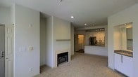 This single-floor space showcases an open-concept design that seamlessly connects the living area to the kitchen. The living room is finished with plush wall-to-wall carpeting and centers around a tile-surround fireplace with a clean mantel shelf, serving as a functional focal point. Recessed lighting throughout provides consistent, even illumination across the neutral-toned walls and white ceilings.

The kitchen is visibly well-appointed, featuring white cabinetry, a mosaic tile backsplash, stainless steel appliances including a refrigerator, and a raised breakfast bar that opens toward the living space. Under-cabinet lighting adds both functionality and ambiance to the kitchen area. Track lighting above the bar area further enhances the workspace.

A doorway visible beyond the kitchen leads to an additional room, suggesting a thoughtful layout that balances open communal areas with private spaces. The overall design palette is neutral and contemporary, with soft grays and whites creating a clean, cohesive aesthetic throughout. The open flow between rooms gives the space a welcoming and practical feel suitable for everyday living.