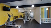 This fitness facility offers a focused and functional workout environment designed to accommodate a range of exercise needs. The space features a bold color scheme, with vibrant yellow accent walls contrasting against crisp white paneled ceilings and dark structural beams, creating an energetic yet clean aesthetic. Globe pendant lights hang from the ceiling, providing warm, even illumination throughout the room. The flooring is finished with durable dark rubberized matting, ideal for gym use and providing both safety and comfort underfoot. A full-length wall mirror spans one side of the room, visually expanding the space and allowing users to monitor their form during workouts. The facility is equipped with a multi-station cable and weight machine at its center, a dumbbell rack stocked with a range of free weights, and cardio equipment including an elliptical trainer. A wall-mounted air conditioning unit ensures a comfortable training environment. An adjacent area is visible through the mirrored reflection, suggesting additional space beyond the main workout floor. Overall, this gym delivers a practical, well-organized setting suitable for both strength training and cardiovascular exercise.