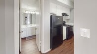 This single-floor residence at 2700 Cottage Pl in Greensboro, North Carolina offers a practical and comfortable layout with a clean, neutral aesthetic throughout. The living areas feature rich hardwood-style flooring that adds warmth and character, while multiple windows dressed with blinds and sheer vertical panels invite abundant natural light and provide views of the surrounding greenery. The kitchen is equipped with white cabinetry, light stone-finish countertops, and black appliances including a refrigerator and range, offering a functional workspace with a classic contrast of tones. The bedrooms are finished with plush carpeting and light gray walls, creating a calm and cozy atmosphere. One bedroom includes a ceiling fan with integrated lighting, while another offers a window with outdoor views. The bathrooms are well-appointed, each featuring a vanity with speckled stone countertops, a large mirror, and bright overhead lighting. One bathroom includes hardwood-style flooring, a shower-tub combination, and ample storage. Throughout the home, textured ceilings, clean white trim, and a consistent soft gray palette contribute to a cohesive and move-in-ready environment.