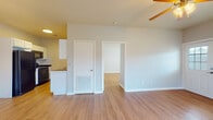 This single-floor residence offers a cohesive and practical layout finished with warm hardwood-style flooring that runs consistently throughout every room, lending a unified and clean aesthetic to the entire space. The open living area is bright and airy, illuminated by natural light and equipped with a ceiling fan, creating a comfortable and well-ventilated environment. A doorway connects the living space directly to additional rooms, ensuring a smooth and intuitive flow throughout the unit. The galley-style kitchen is efficiently designed, featuring white cabinetry on both sides, dark countertops, a double sink, and black appliances including a full-size refrigerator and dishwasher. Ample upper and lower cabinet storage makes this kitchen both functional and organized. The bedrooms are generously proportioned, each featuring ceiling fans with integrated lighting, windows dressed with blinds for natural light and privacy, and closet access. The neutral gray walls provide a versatile backdrop throughout the home. The bathroom is notably spacious, offering a long dual-sink vanity with white cabinetry, mirrored medicine cabinets, and a separate water closet, all finished with the same warm flooring. The overall ambiance of this residence is clean, neutral, and move-in ready.