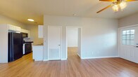 This single-floor residence offers a cohesive and practical layout finished with warm hardwood-style flooring that runs consistently throughout every room, lending a unified and clean aesthetic to the entire space. The open living area is bright and airy, illuminated by natural light and equipped with a ceiling fan, creating a comfortable and well-ventilated environment. A doorway connects the living space directly to additional rooms, ensuring a smooth and intuitive flow throughout the unit. The galley-style kitchen is efficiently designed, featuring white cabinetry on both sides, dark countertops, a double sink, and black appliances including a full-size refrigerator and dishwasher. Ample upper and lower cabinet storage makes this kitchen both functional and organized. The bedrooms are generously proportioned, each featuring ceiling fans with integrated lighting, windows dressed with blinds for natural light and privacy, and closet access. The neutral gray walls provide a versatile backdrop throughout the home. The bathroom is notably spacious, offering a long dual-sink vanity with white cabinetry, mirrored medicine cabinets, and a separate water closet, all finished with the same warm flooring. The overall ambiance of this residence is clean, neutral, and move-in ready.