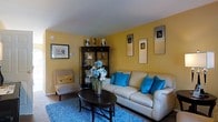 This multi-floor space spans three levels, offering a generous layout with distinct living areas across each floor. The living room is immediately welcoming, characterized by warm golden-yellow walls that create an energetic yet cozy atmosphere. A cream-toned sofa anchors the seating area, accented by vivid turquoise throw pillows that provide a striking contrast against the warm wall color. A plush blue shag rug defines the conversation area, adding texture and comfort underfoot. A dark wood oval coffee table serves as a functional centerpiece, topped with a decorative floral arrangement featuring blue and white blooms. A matching dark wood display cabinet in the corner provides both storage and a showcase for decorative objects. Wall-mounted framed artwork adds a personalized touch to the space. A neutral armchair with a coordinating cushion completes the seating arrangement, offering additional comfort. The room is finished with standard ceiling height and benefits from ambient lighting. The overall aesthetic balances bold color choices with neutral furnishings, creating a lively yet cohesive interior environment across this thoughtfully arranged multi-floor property.