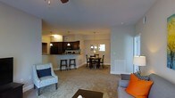 This single-floor residence showcases an open-concept layout that effortlessly connects the living, dining, and kitchen areas into one cohesive and functional space. The living room is generously proportioned, featuring plush carpeted flooring, a ceiling fan, and ample room for comfortable seating arrangements. A large window along one wall invites natural light, brightening the neutral-toned interior.

The kitchen is defined by dark cabinetry that contrasts elegantly with light countertops, and is equipped with full-sized appliances including a refrigerator and range. A raised breakfast bar with barstool seating extends from the kitchen counter, creating a casual dining option while maintaining an open sightline to the rest of the living space.

Adjacent to the kitchen, the dedicated dining area accommodates a full dining table and chairs beneath a classic chandelier, adding a refined touch to everyday meals. A window in the dining area further enhances the natural light throughout.

The consistent carpeted flooring ties the entire space together, while the neutral wall palette and warm lighting create an inviting and comfortable ambiance. The thoughtful layout maximizes the sense of openness, making this a welcoming space well-suited for both relaxed daily living and entertaining.