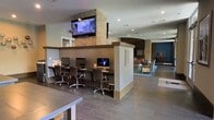 This two-story space offers a well-appointed communal environment designed with both productivity and relaxation in mind. The main level showcases a built-in business center equipped with multiple desktop computers and ergonomic task chairs, set along a dedicated workstation counter — ideal for those needing a professional workspace. A wall-mounted flat-screen television is positioned above the workstation area, anchored by a striking exposed brick column that adds a warm, textural contrast to the otherwise contemporary interior. The flooring throughout features large-format tile in a rich wood-tone finish, lending a clean and cohesive look to the space. Adjacent to the business center, a comfortable lounge area provides casual seating arrangements with sofas and accent chairs set atop an area rug, creating an inviting retreat. Recessed lighting illuminates the space evenly, complemented by a neutral color palette of warm grays and whites. A bold teal accent wall in the lounge area adds a pop of color, while artwork and decorative elements contribute to the modern, community-focused atmosphere. The open-plan layout encourages flow between functional zones, making this a versatile and welcoming environment throughout both floors.