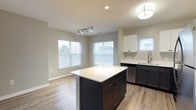 This 2-bedroom, 2-bathroom residence offers a thoughtfully designed single-floor layout with contemporary finishes throughout. The open-concept kitchen and living area serves as the heart of the home, featuring warm wood-look flooring, abundant natural light from multiple windows, and sliding glass doors that open to a private balcony overlooking the outdoors. The kitchen is equipped with stainless steel appliances — including a gas range, built-in microwave, dishwasher, and refrigerator — complemented by a two-tone cabinetry scheme of white upper cabinets and dark lower cabinets, crisp white countertops, and a central island ideal for casual dining or meal preparation. The living area provides generous space with a clean, neutral palette and easy access to the entry hallway. Both bedrooms are carpeted for comfort and feature windows that bring in natural light. The primary bedroom includes access to a walk-in closet and an en-suite bath. The two bathrooms are finished with white tile, chrome fixtures, and dark-toned vanities. Throughout the residence, neutral gray walls and clean white trim create a modern, cohesive aesthetic that is both functional and inviting.